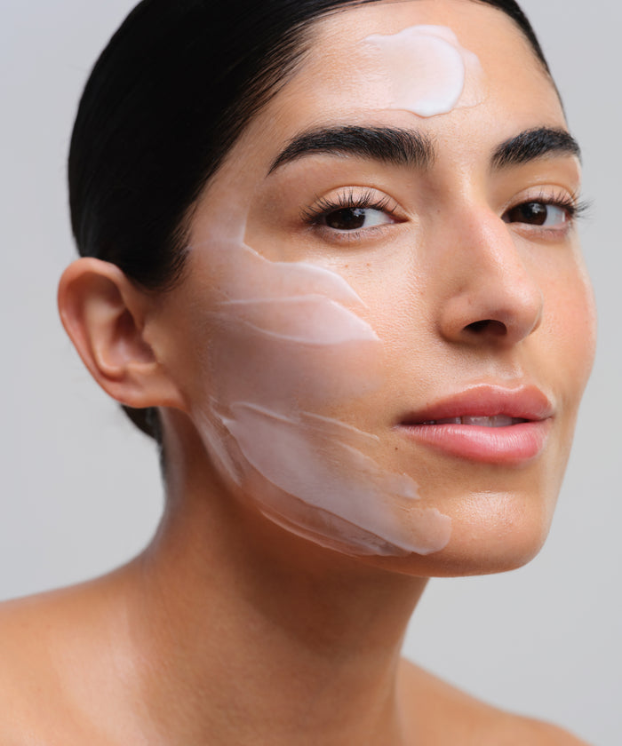 A woman with smooth, sensitive skin and dark hair pulled back has Nourishing Cleansing Milk applied to her forehead and cheeks, confidently facing the camera against a plain light background.