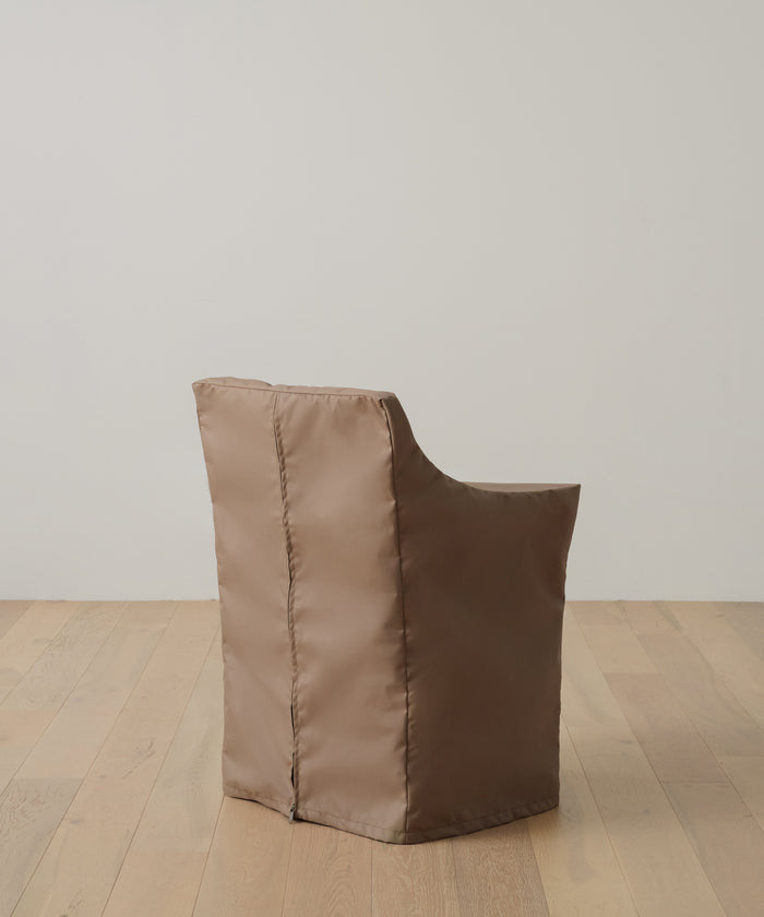 A brown, padded chair with a shiny fabric cover sits on a light wood floor by a white wall. Protect it and keep it looking new with the Outdoor Montecito Dining Arm Chair Cover, designed for a perfect fit and lasting durability.