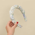 A hand with red nail polish holds a padded fabric headband featuring a white background with a delicate blue, green, and pink floral vine pattern. A white fabric tag is visible on the inside of the headband, reading 'DAMASK HEADBANDS'.