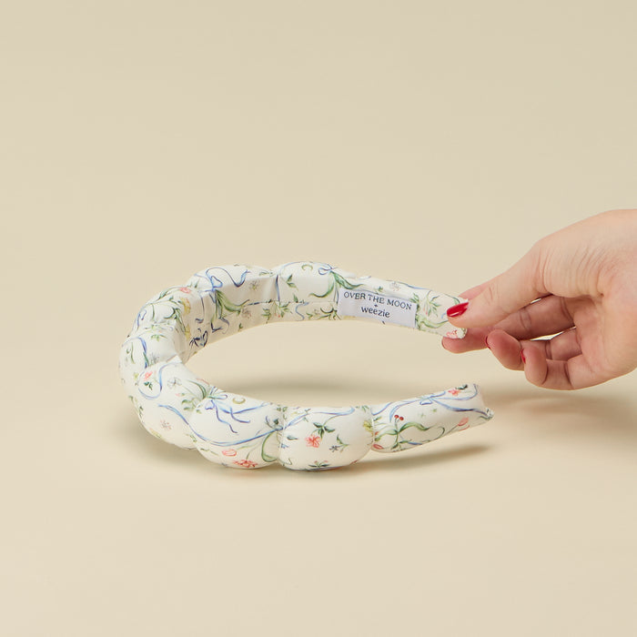 A hand with red nail polish holding up one end of a padded, braided fabric bubble headband to reveal an interior tag that reads "OVER THE MOON + weezie". The headband features a white background with a delicate blue ribbon, green vine, and pink floral pattern, set against a plain beige background.