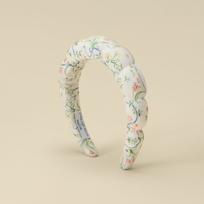 A twisted fabric headband with a delicate floral pattern in soft pink, green, and blue on an off-white background, displayed on a solid beige studio background.