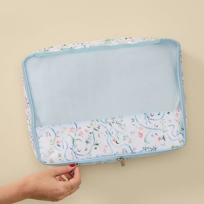 A light blue and white floral mesh zippered travel organizer pouch being held by a hand against a solid beige background.