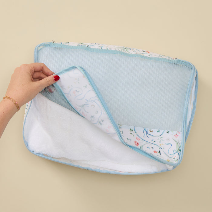 A hand with red nail polish lifts the corner of a light blue mesh travel organizer pouch to reveal a white and floral patterned lining, set against a solid beige background.