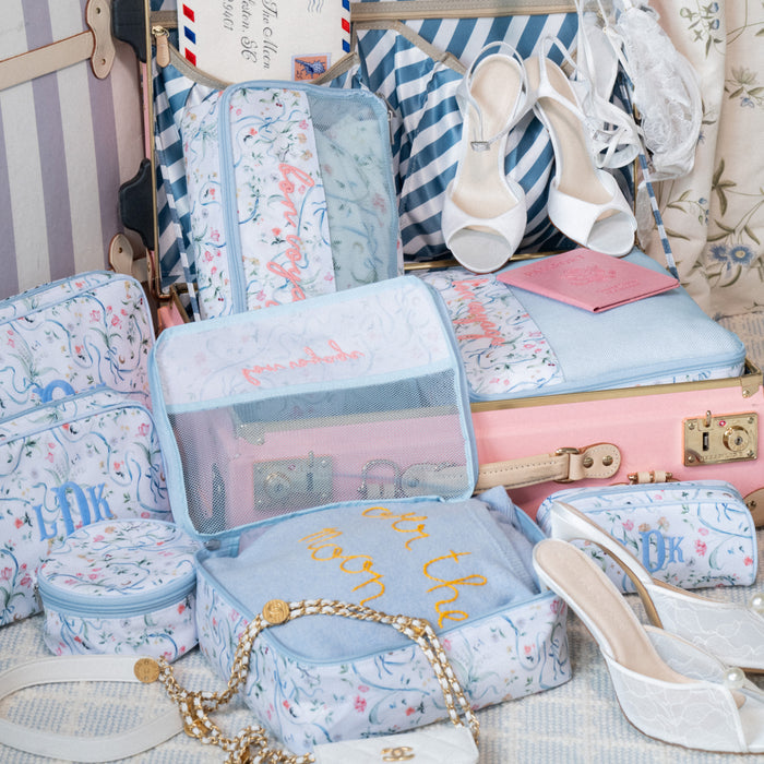 A coordinated set of pastel travel packing cubes and small zip pouches with a light blue floral print, arranged around a pink suitcase. White heeled sandals, a striped bag, and other travel accessories are visible in the styled scene.