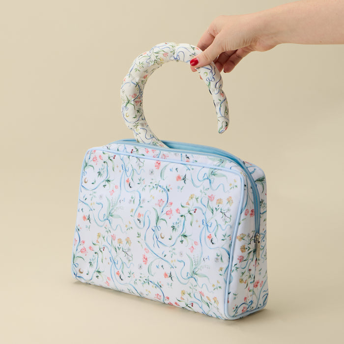 A light blue and white floral patterned cosmetic bag with a curved top handle, held by a hand with red nail polish against a solid beige background.