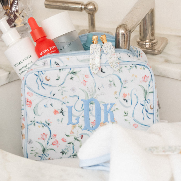 A floral toiletry bag with blue monogrammed initials 'LDK' sits in a bathroom sink next to skincare bottles, with a marble countertop and chrome faucet visible.
