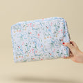 A hand holding a rectangular zippered toiletry bag with a white background and a delicate blue, pink, and green floral pattern, set against a solid beige background.