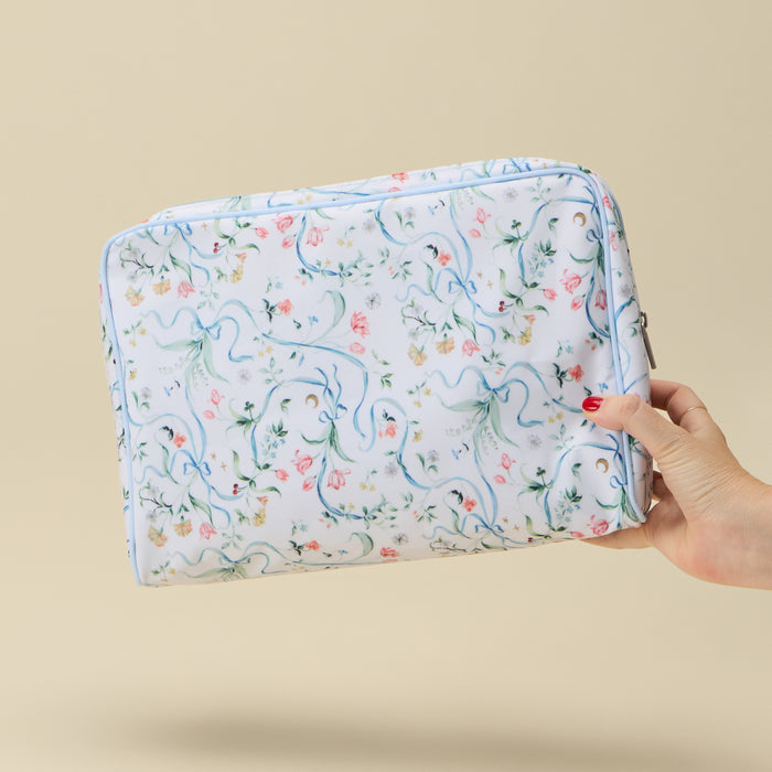 A hand holding a rectangular zippered toiletry bag with a white background and a delicate blue, pink, and green floral pattern, set against a solid beige background.