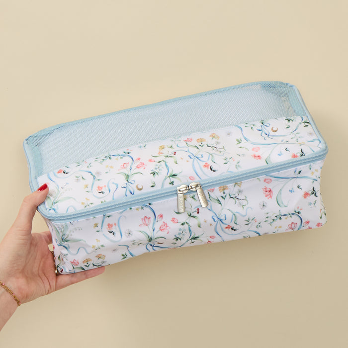 A hand holding a rectangular zippered pouch with a white background, a delicate multicolor floral and vine print, and a light blue mesh top and zipper trim, set against a solid beige background.