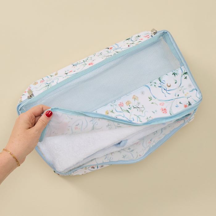 A hand with red nail polish holds open a light blue mesh zippered organizer bag with a white floral fabric exterior, showing a white textured insert inside, set against a solid beige background.