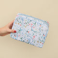 A hand with red nail polish holds a rectangular zippered pouch featuring a delicate floral and scroll pattern in light blue, pink, green, and yellow on a white background, set against a solid beige backdrop.