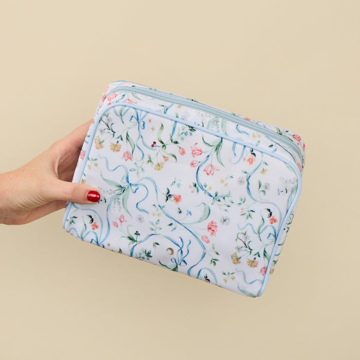 A hand with red nail polish holds a rectangular zippered pouch featuring a delicate floral and scroll pattern in light blue, pink, green, and yellow on a white background, set against a solid beige backdrop.