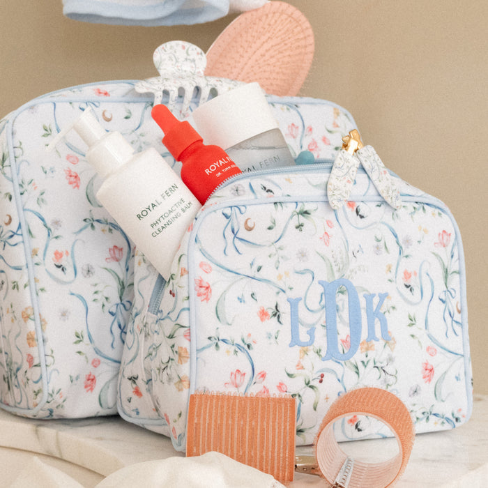 A floral toiletry bag with monogrammed initials 'LDR' in light blue, styled with skincare items including a white bottle labeled Royal Cashmere and a red dropper bottle, on a neutral beige background.