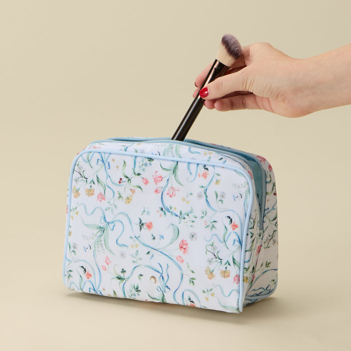 A hand with red nail polish places a black-handled makeup brush into a partially open, medium-sized toiletry bag. The bag features a white background with a delicate pattern of blue ribbons, green vines, and small pink and yellow flowers, accented by light blue piping, and is set against a plain beige background.