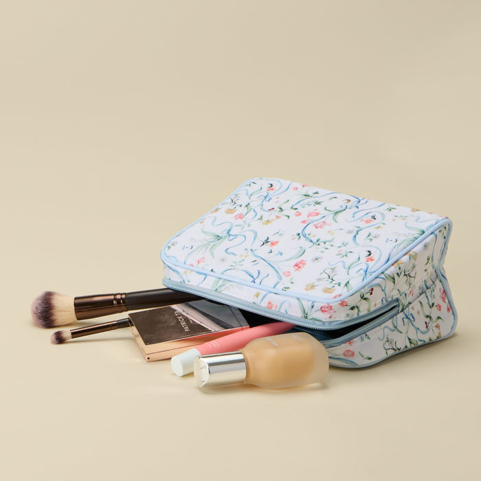 A rectangular makeup bag with a light blue and white floral pattern, shown with makeup items like brushes, a compact, and a foundation bottle spilling out from the front zipper. The bag is photographed on a solid beige background.