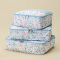 Three stacked zippered travel packing cubes with a white background and a delicate pastel floral and vine pattern, featuring light blue trim and silver zippers, set against a solid beige background.