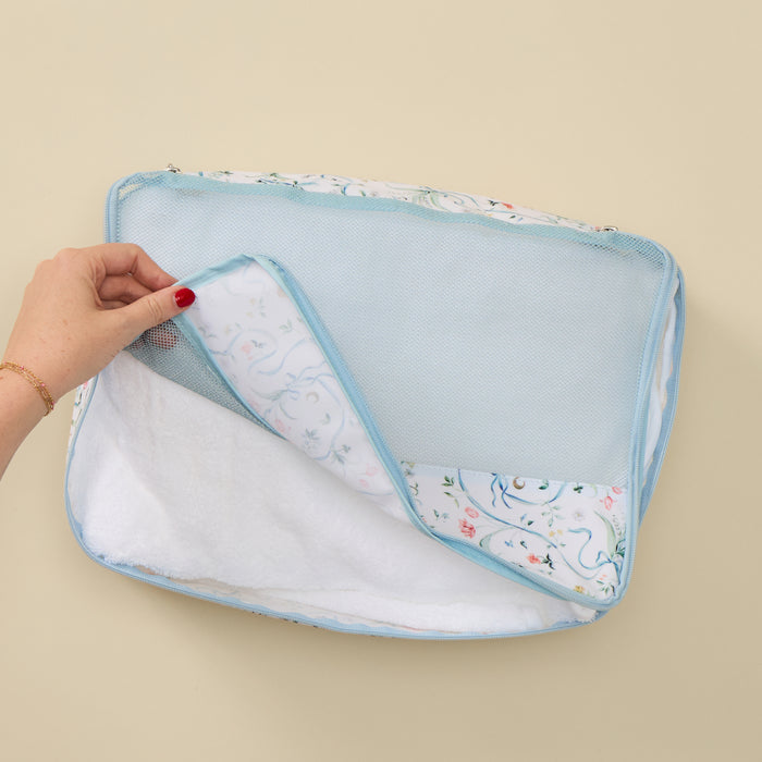 A hand with red nail polish lifts the corner of a light blue mesh organizer bag to reveal a white fabric insert with a delicate pastel floral pattern, set against a solid beige background.