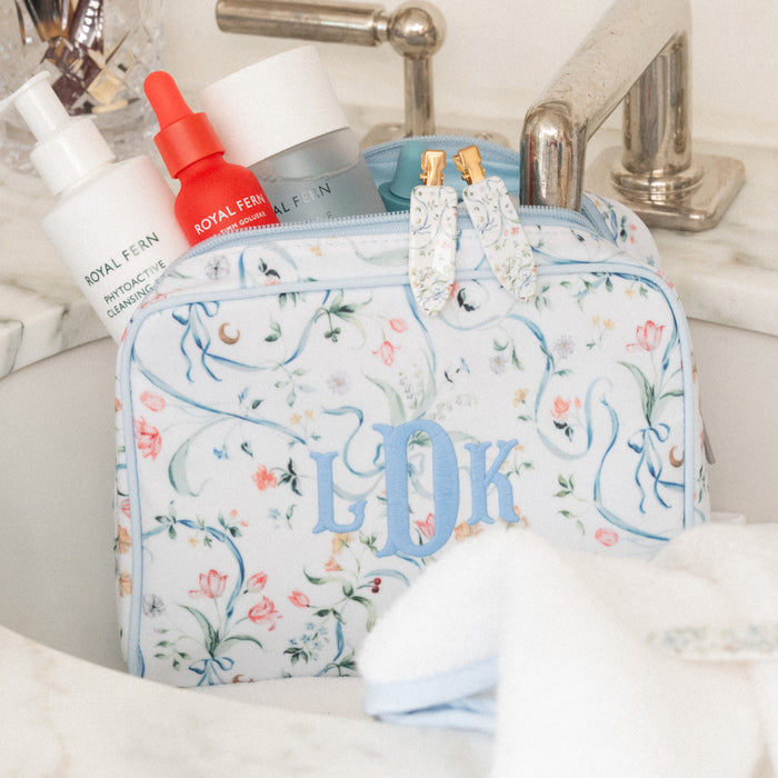 A floral toiletry bag with blue monogrammed initials 'L&K' sits in a bathroom sink next to Royal Fern skincare bottles. The bag features a delicate blue and pink floral pattern on a white background with gold zipper pulls.