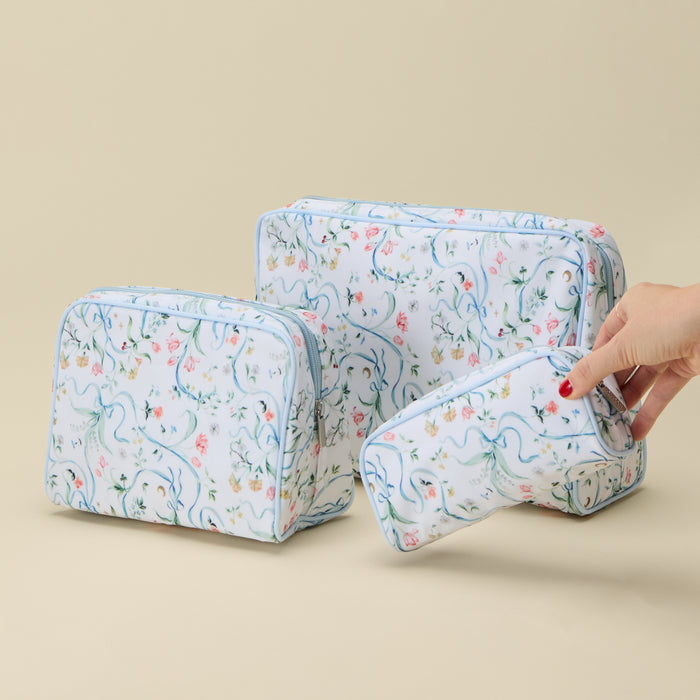 Three floral toiletry bags in different sizes with a light blue border and a delicate multicolor print, shown on a beige background with a hand holding the smallest bag.