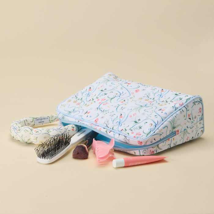 A fabric makeup bag with a light blue and white floral pattern, shown with various beauty accessories like a hairbrush, lip balm, and a small pink item on a solid beige background.
