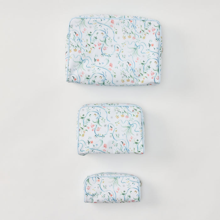 A set of three toiletry bags in small, medium, and large sizes arranged vertically on a plain white background. The bags feature a delicate pattern of blue ribbons, green vines, and small pink and yellow flowers, accented by light blue piping.