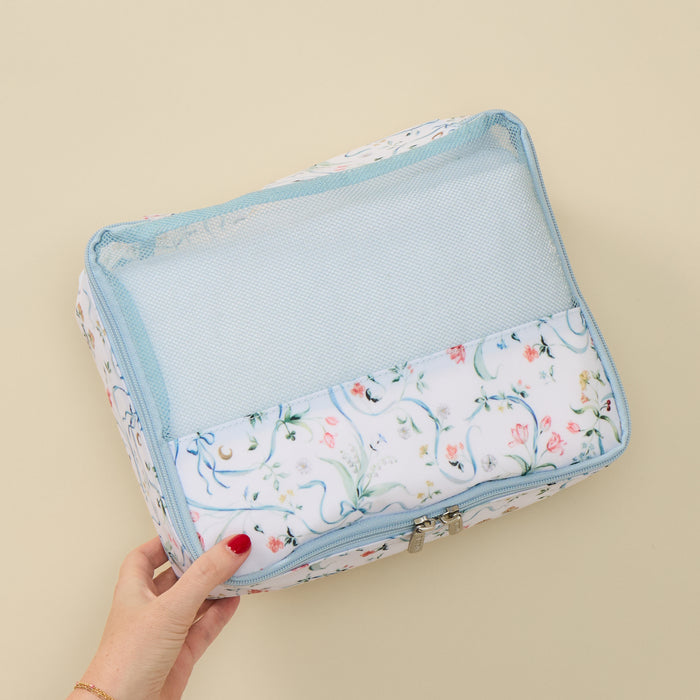 A hand with red painted nails holds a rectangular travel organizer bag with a floral print and light blue mesh pocket, set against a solid beige background.