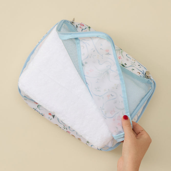A hand with red nail polish pulling back the flap of a small packing cube to reveal a folded white towel inside. The packing cube features a delicate pattern of blue ribbons, green vines, and small pink flowers on a white background, accented by light blue piping and a light blue mesh top panel, set against a plain beige background.