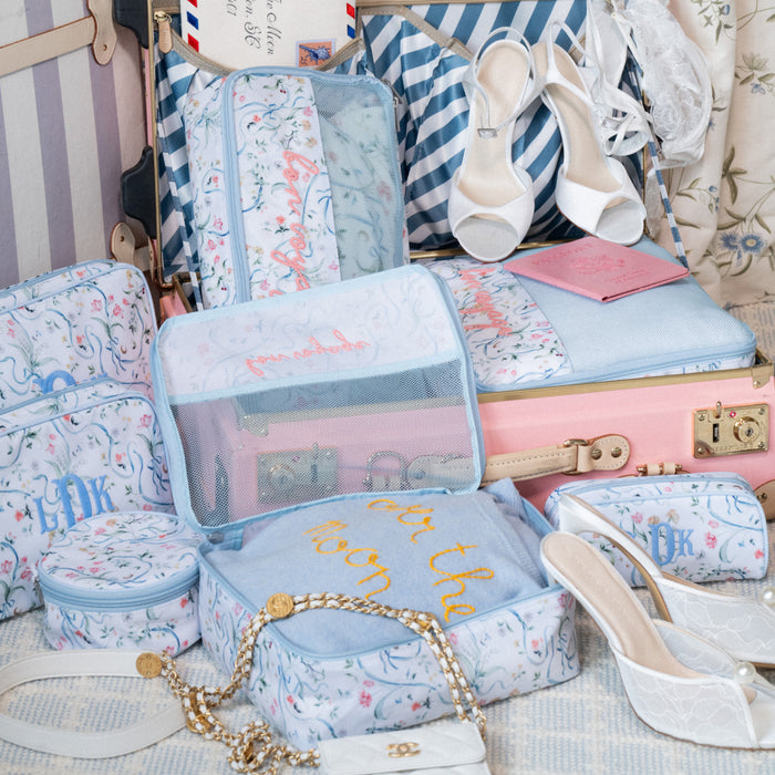A styled travel collection featuring a light pink vintage suitcase overflowing with matching packing cubes and toiletry bags. The bags feature a delicate pattern of blue ribbons, green vines, and small pink flowers, accented by light blue piping and mesh tops. Several pieces display custom embroidery in red, yellow, and blue, including phrases like "bon voyage" and monograms.