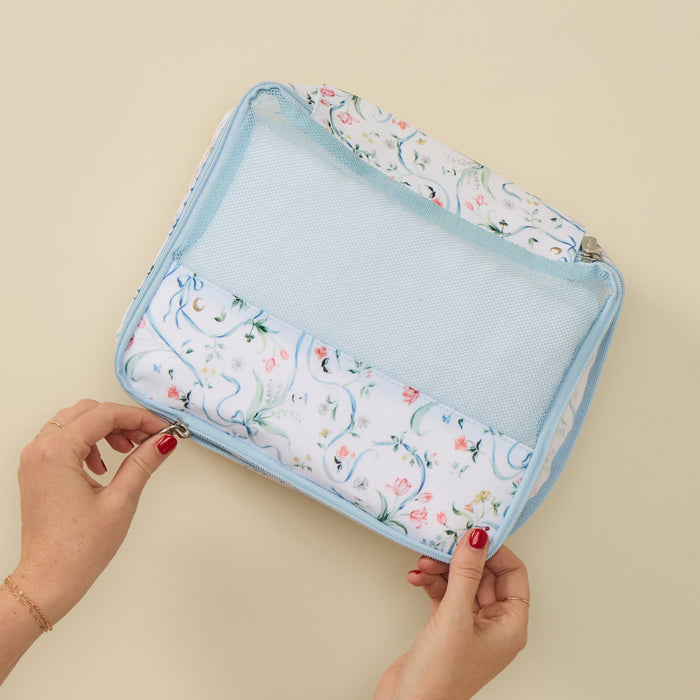 A light blue mesh zippered travel organizer bag with a white floral print fabric lining, held open by two hands with red nail polish against a solid beige background.