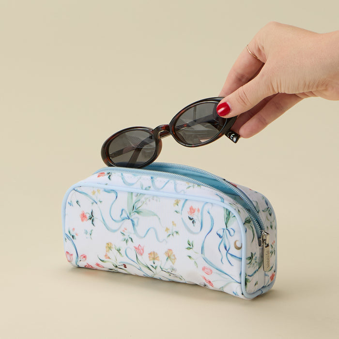 A hand with red nail polish holds a pair of dark sunglasses above a zippered cosmetic pouch. The pouch has a white background with a delicate pastel floral print and light blue wavy line details, set against a solid beige background.