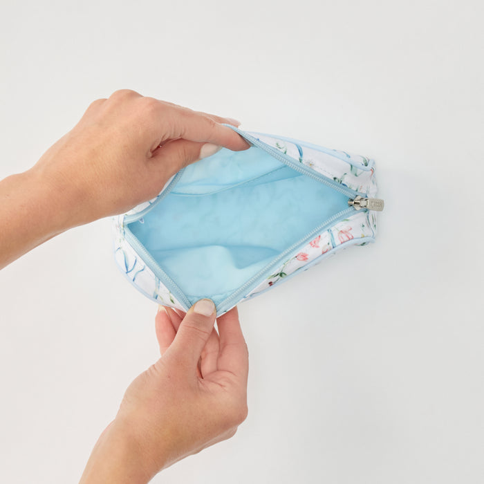 A close-up of a person's hands holding open a zippered cosmetic pouch. The pouch has a white fabric exterior with a delicate pink and green floral print, and a light blue interior lining.