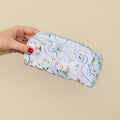 A hand with red nail polish holds a white zippered pencil case decorated with a delicate blue, pink, and yellow floral pattern against a solid beige background.