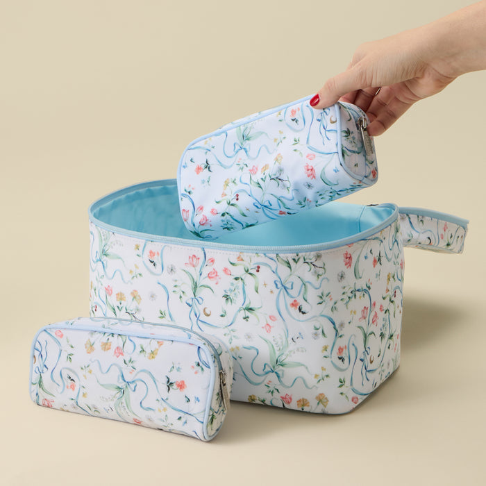 Three floral zip pouches in different sizes with a light blue lining, featuring a delicate watercolor-style print of pink flowers, green leaves, and blue vines. A hand is shown holding the largest pouch, set against a solid beige background.