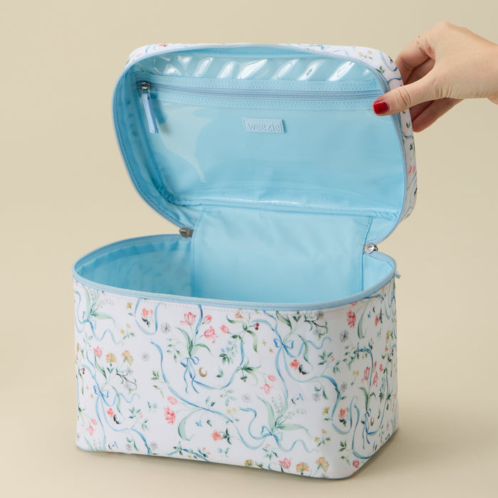 A rectangular cosmetic bag with a white floral print exterior and a light blue interior, shown open with a hand lifting the lid. The brand name 'woosie' is visible on a small interior label.