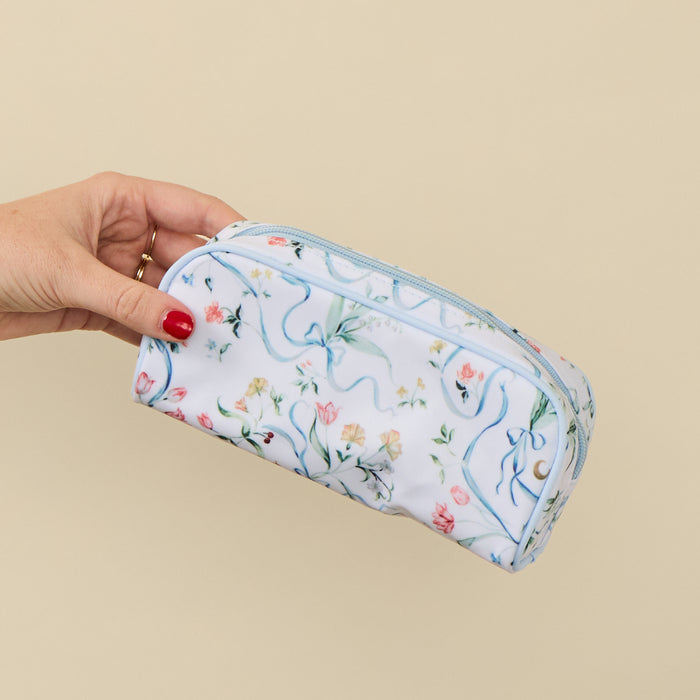 A hand with red nail polish holds a white zippered pencil case decorated with a delicate blue, pink, and green floral and vine pattern against a solid beige background.