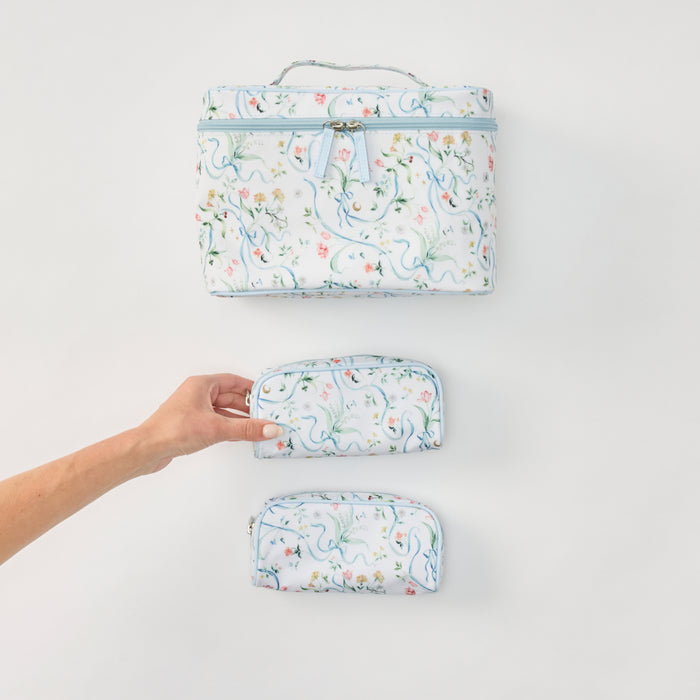 Three matching fabric zippered pouches or small cases with a white background and a delicate multicolor floral print, arranged vertically on a light gray surface. A human hand is holding the middle case.