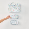 Three matching fabric zippered pouches or small cases with a white background and a delicate multicolor floral print, arranged vertically on a light gray surface. A human hand is holding the middle case.