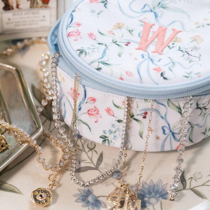 A round fabric jewelry organizer pouch with a floral print and embroidered monogram 'W', styled with gold and silver chain necklaces draped over it on a matching floral surface.