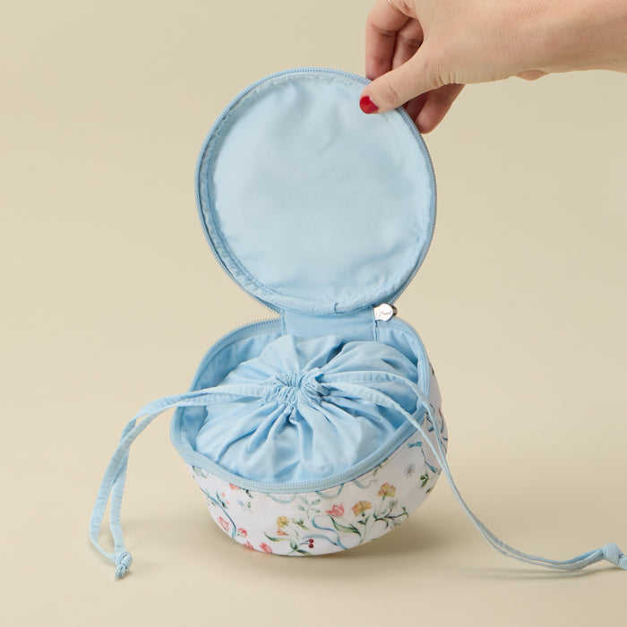 A hand with red nail polish holds open a round drawstring bag with a light blue interior and a white floral exterior, set against a solid beige background.