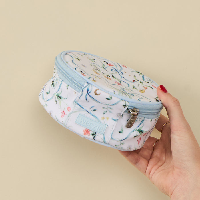 A hand with red nail polish holds a small zippered pouch against a solid beige background. The pouch is white with a light blue zipper trim and features a whimsical print of a blue cat, a crescent moon, and small pink and yellow flowers.