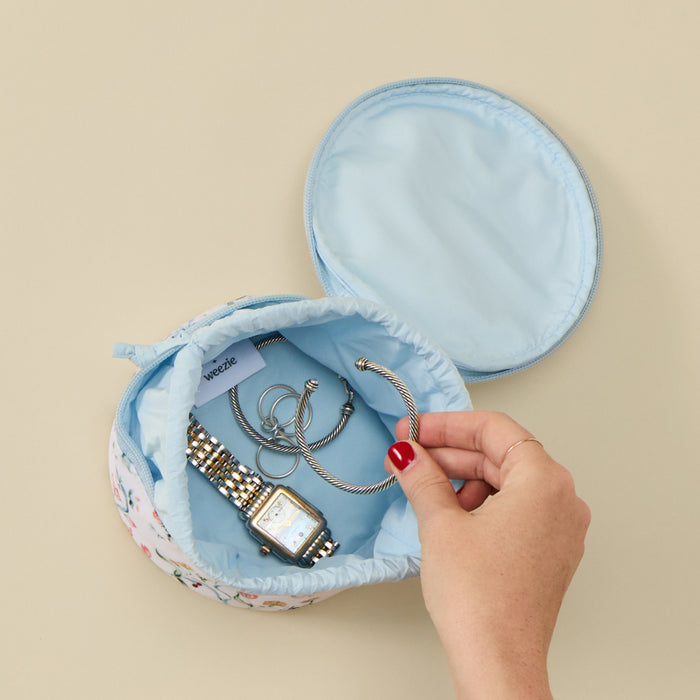 A light blue fabric jewelry organizer pouch with a floral patterned exterior, shown open with jewelry inside and a hand reaching in. The pouch is photographed on a plain beige background.