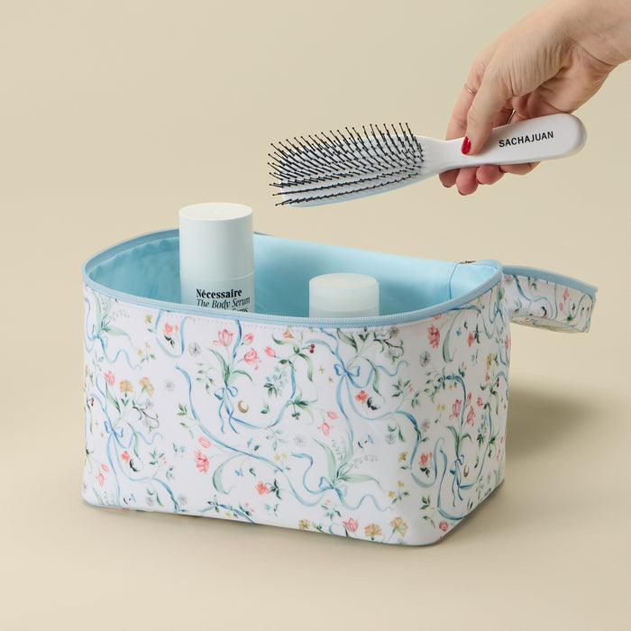A floral toiletry bag with a light blue interior sits on a beige background. A hand holds a white hairbrush above the bag, which contains two white bottles.