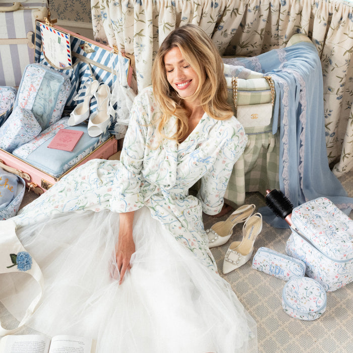 A woman sits on a patterned floor surrounded by matching travel and toiletry bags, a suitcase, and a bed in a styled bedroom setting. She is wearing a white tulle robe with a light blue and pink floral print.