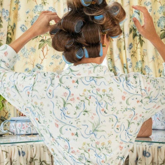 A woman viewed from behind adjusting large blue hair rollers, wearing a white lightweight robe with a blue, green, and pink floral ribbon print. She is seated at a glass vanity table in front of curtains that feature the identical matching floral pattern.