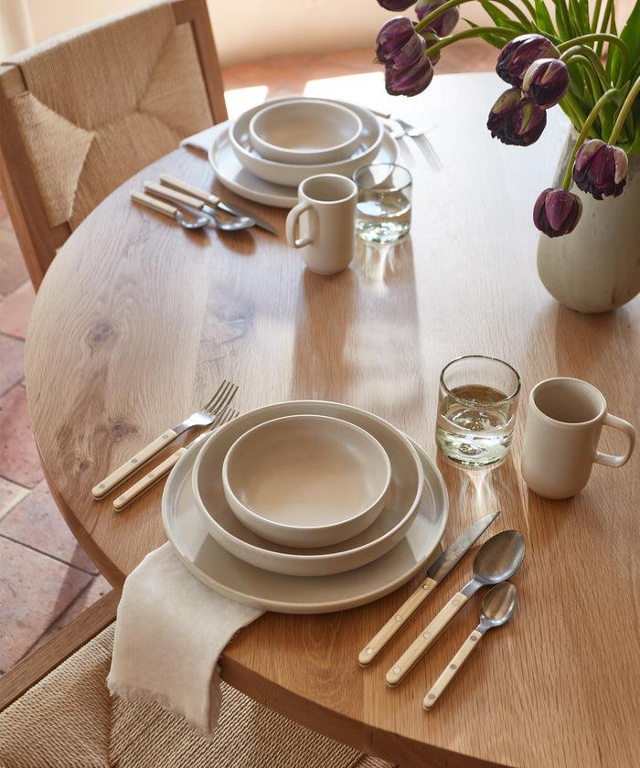 A round wooden table set for two with Pacific Breakfast Bowls, white stoneware mugs, and glasses of water. A vase of purple tulips decorates the scene as sunlight streams in, creating a warm, inviting atmosphere.
