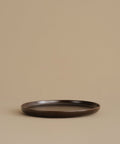 A dark brown ceramic dinner plate with a matte finish, photographed against a solid beige background.