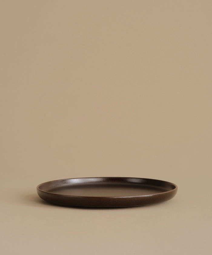 A dark brown ceramic dinner plate with a matte finish, photographed against a solid beige background.
