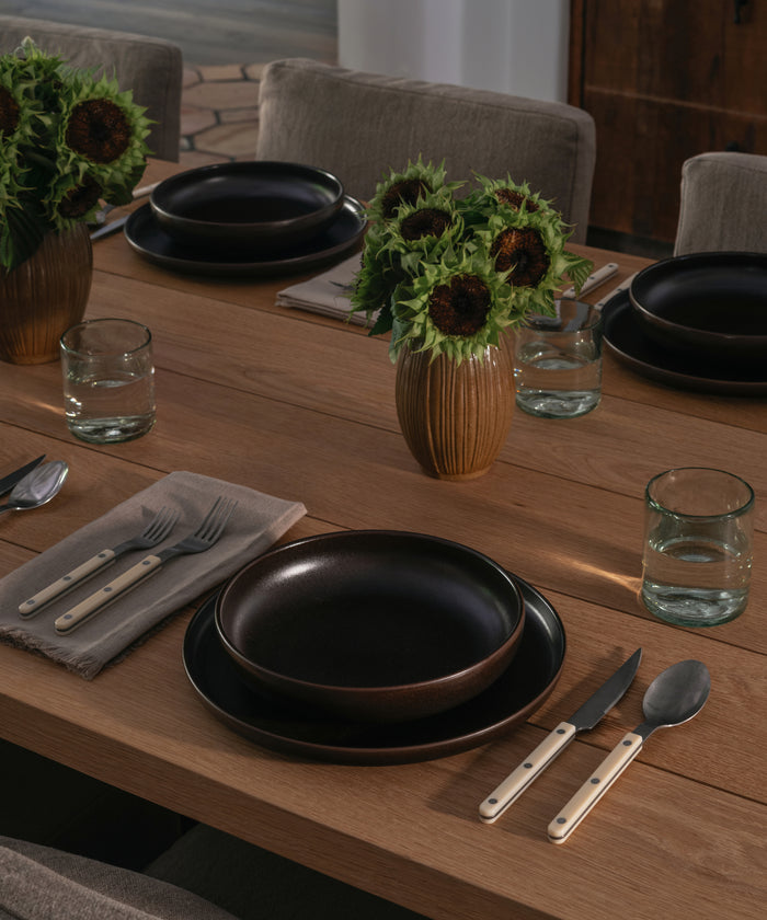 A styled dining table setting with matte dark brown ceramic plates, matching black bowls, clear glass water glasses, silver cutlery with light wood handles, and two ribbed vases filled with sunflower arrangements on a wooden table.