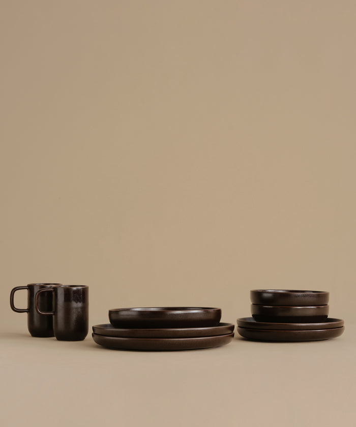 Dark brown ceramic tableware set including plates, bowls, and mugs, arranged on a plain beige studio background.