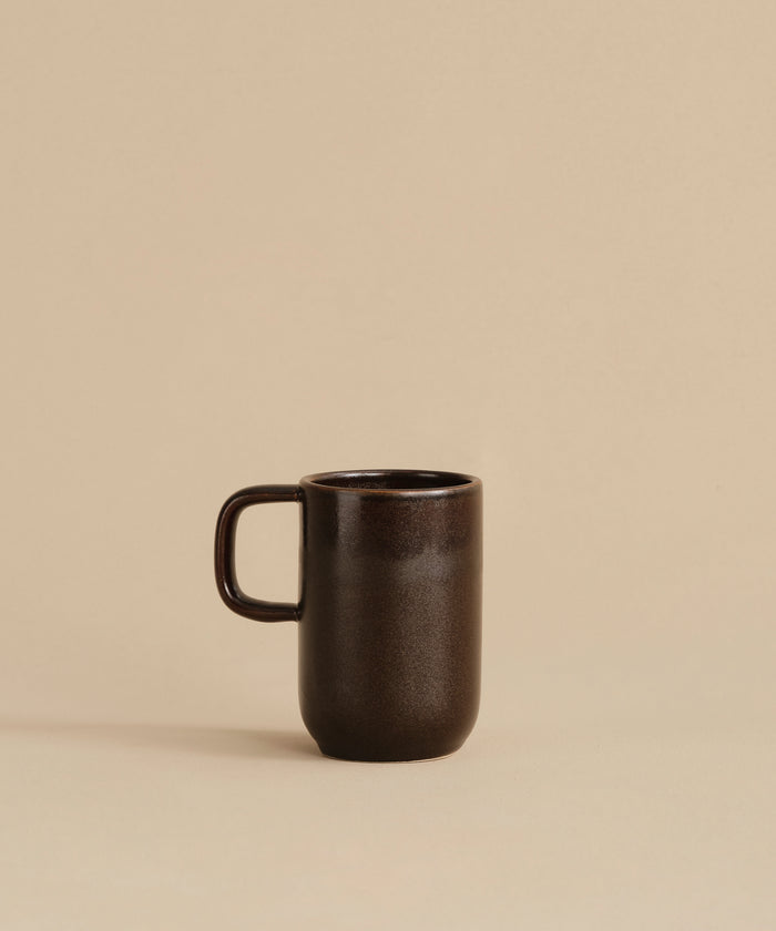 The Pacific Mug, made of dark chocolate-colored stoneware with a handle, rests on a beige surface against a plain beige background.
